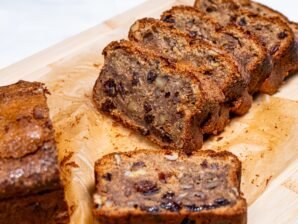 A loaf of banana bread sliced into thick pieces, showing a moist crumb filled with walnuts and raisins on a wooden cutting board.