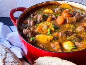 Guinness beef stew in a red Dutch oven with chunks of beef, carrots, corn, peas, and potatoes in a rich sauce, served with crusty bread.