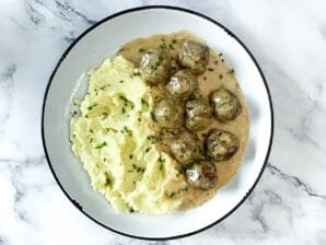 A plate of creamy meatballs in rich sauce, served with fluffy mashed potatoes and garnished with fresh chives, on a marble background.