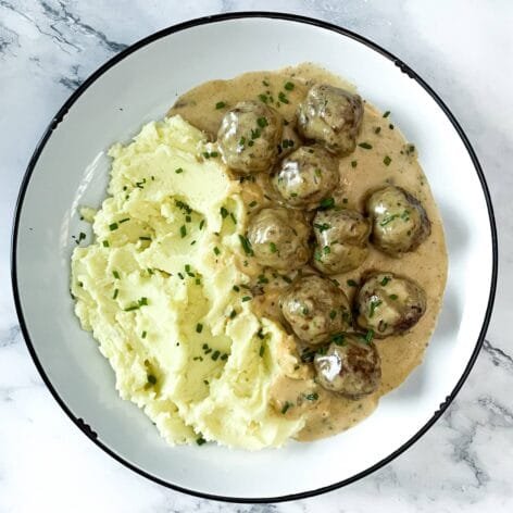 A plate of creamy meatballs in rich sauce, served with fluffy mashed potatoes and garnished with fresh chives, on a marble background.