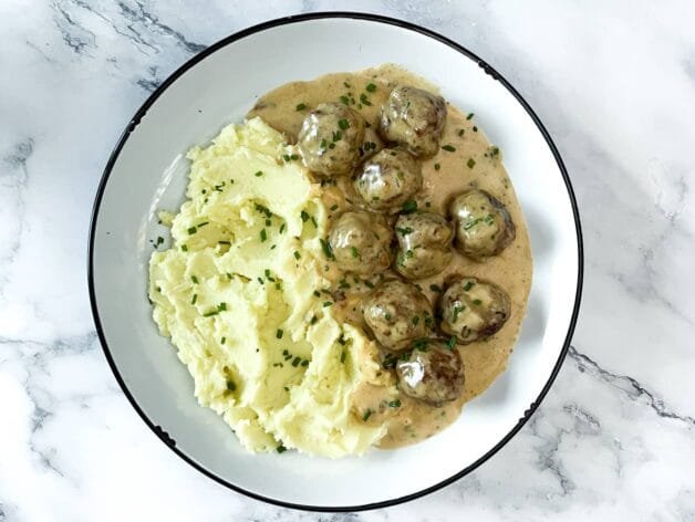 A plate of creamy meatballs in rich sauce, served with fluffy mashed potatoes and garnished with fresh chives, on a marble background.