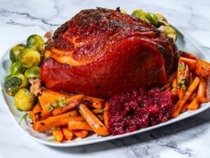 Honey-Dijon glazed ham served with roasted carrots, Brussels sprouts, and cranberry sauce on a white platter.