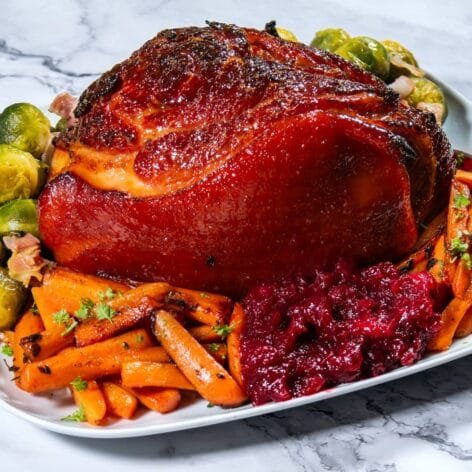 Honey-Dijon glazed ham served with roasted carrots, Brussels sprouts, and cranberry sauce on a white platter.