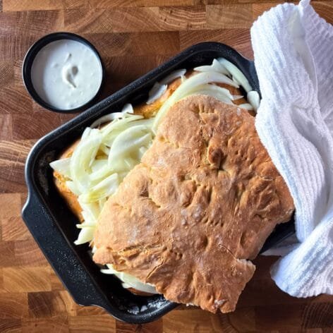 Freshly baked savory onion flatbread in a black baking pan, topped with salted onions, partially covered with a white kitchen towel, and served with a side of sour cream.