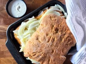 Freshly baked savory onion flatbread in a black baking pan, topped with salted onions, partially covered with a white kitchen towel, and served with a side of sour cream.