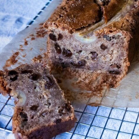 A sliced loaf of banana bread showing a moist crumb with walnuts, raisins, and caramelized banana topping on a cooling rack.