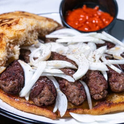 Open-faced cevapi sandwich topped with sliced onions, served with a side of ajvar.