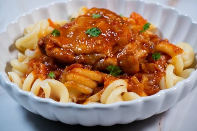 Close-up of chicken in tomato sauce served over pasta, garnished with parsley.