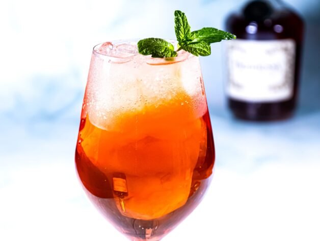 A Citrus Hennessy Spritz cocktail served in a tall glass with fresh mint garnish, with a Hennessy bottle blurred in the background.