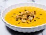 A bowl of creamy butternut squash soup, topped with golden croutons, toasted pumpkin seeds, and a drizzle of olive oil, served in a white scalloped bowl.
