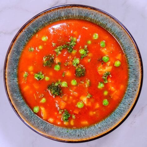 A vibrant bowl of hearty tomato vegetable soup garnished with broccoli, peas, and fresh parsley, served in a rustic blue ceramic dish.