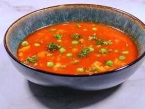A side view of a bowl filled with hearty tomato vegetable soup, garnished with green peas, broccoli, and fresh parsley.