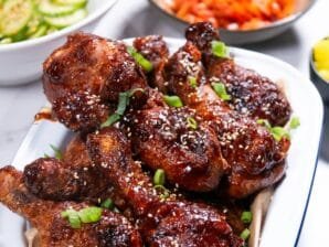 Korean fried chicken drumsticks coated in sweet-and-spicy Yangnyeom sauce, garnished with sesame seeds and green onions, served on a white platter.