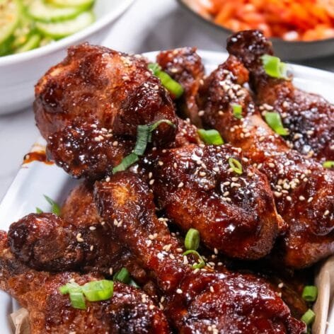 Korean fried chicken drumsticks coated in sweet-and-spicy Yangnyeom sauce, garnished with sesame seeds and green onions, served on a white platter.