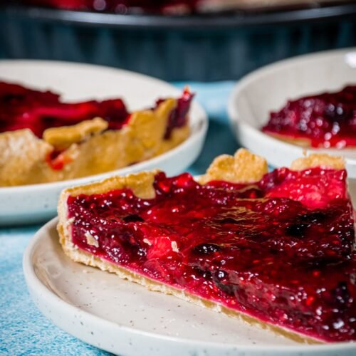 A slice of homemade mixed berry pie with a golden shortcrust pastry and glossy, juicy berry filling, served on a ceramic plate.