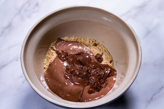 Melted chocolate and Nutella poured over ground nuts and biscuits in a beige bowl.