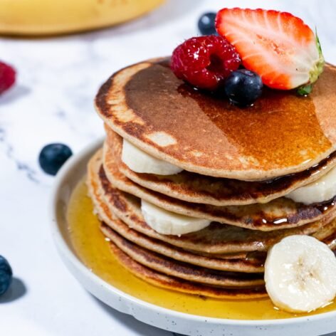 Stack of oatmeal pancakes topped with fresh fruit and maple syrup on a marble surface.