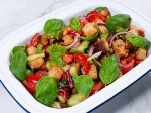 Panzanella salad with roasted bell peppers, crispy bread cubes, fresh basil, and vegetables in a white serving dish.