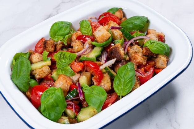 Panzanella salad with roasted bell peppers, crispy bread cubes, fresh basil, and vegetables in a white serving dish.