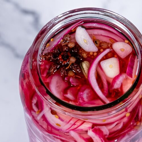 Open jar of pickled red onions showing vibrant pink onion slices, garlic, and spices.