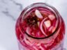 Open jar of pickled red onions showing vibrant pink onion slices, garlic, and spices.