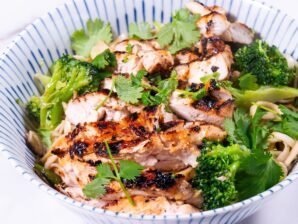 Close-up of stir-fried noodles, broccoli, and grilled chicken garnished with fresh cilantro.