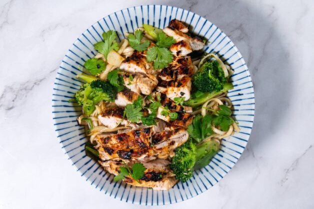 A vibrant bowl of stir-fried Shandon noodles, broccoli, and grilled chicken topped with fresh cilantro.