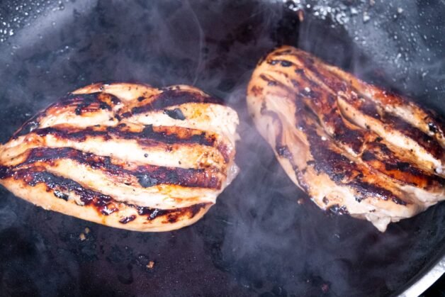 Scored chicken breasts searing in a pan with smoky grill marks.
