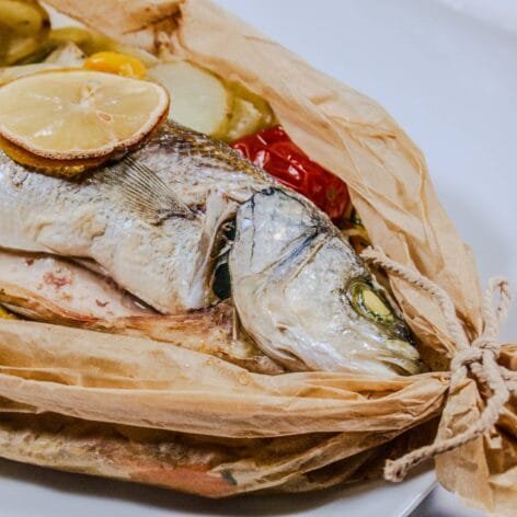 Sea Bass en Papillote with lemon slices, cherry tomatoes, and vegetables in parchment paper on a white plate.