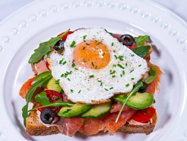 Smoked salmon and avocado toast topped with a sunny-side-up egg, garnished with fresh chives on a white plate.