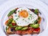Smoked salmon and avocado toast topped with a sunny-side-up egg, garnished with fresh chives on a white plate.