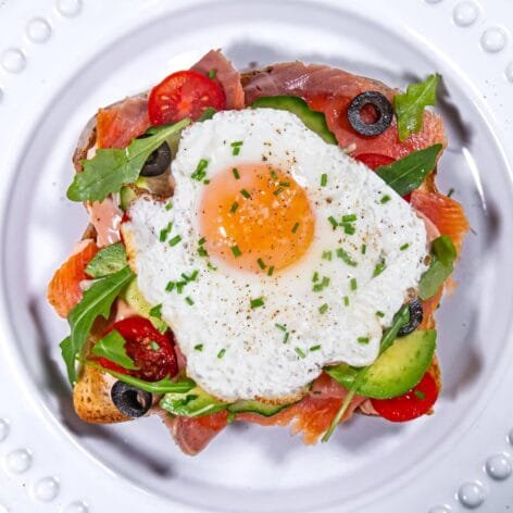 Top-down view of smoked salmon and avocado toast topped with a sunny-side-up egg, surrounded by vibrant veggies and garnished with fresh chives.