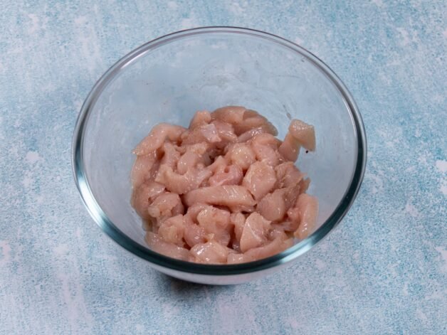 Raw chicken strips marinated in a glass bowl with spices, Shaoxing wine, and cornstarch.