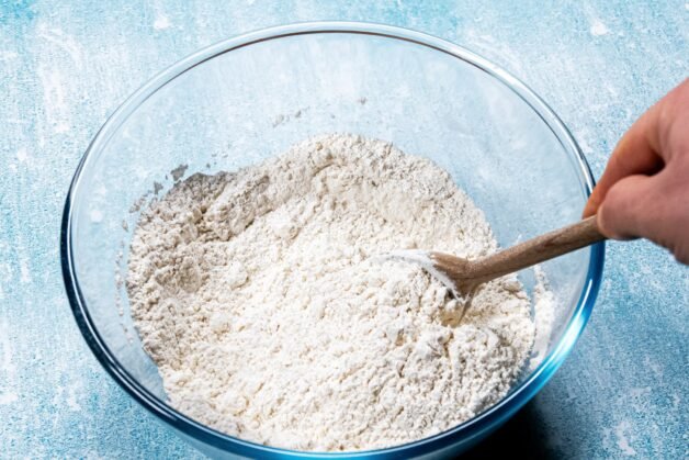 A glass bowl filled with flour, yeast, sugar, and salt being mixed with a wooden spoon.