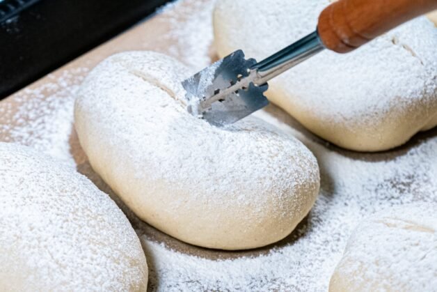 A hand scoring the dusted buns with a sharp razor for a decorative finish.