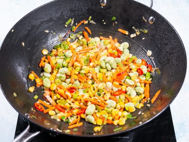 A mix of colorful vegetables, including carrots, broad beans, and sweet corn, stir-frying in a wok.