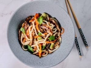 A bowl of udon noodle stir-fry with ground pork, shiitake mushrooms, bell peppers, and cilantro garnish, served with chopsticks on a marble surface.
