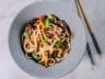 A bowl of udon noodle stir-fry with ground pork, shiitake mushrooms, bell peppers, and cilantro garnish, served with chopsticks on a marble surface.