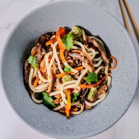A bowl of udon noodle stir-fry with ground pork, shiitake mushrooms, bell peppers, and cilantro garnish, served with chopsticks on a marble surface.
