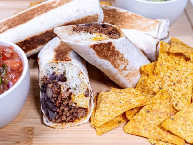 Close-up of a homemade beef burrito cut in half, showing seasoned beef, black beans, cilantro lime rice, and melted cheese, served with tortilla chips.