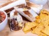 Close-up of a homemade beef burrito cut in half, showing seasoned beef, black beans, cilantro lime rice, and melted cheese, served with tortilla chips.