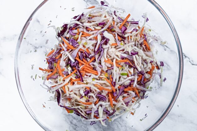 A glass bowl filled with shredded green and red cabbage, carrots, and chopped green onions for coleslaw salad.