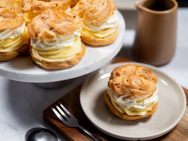 Golden, crispy cream puffs filled with rich vanilla pastry cream and fluffy whipped cream, elegantly served on a marble cake stand.