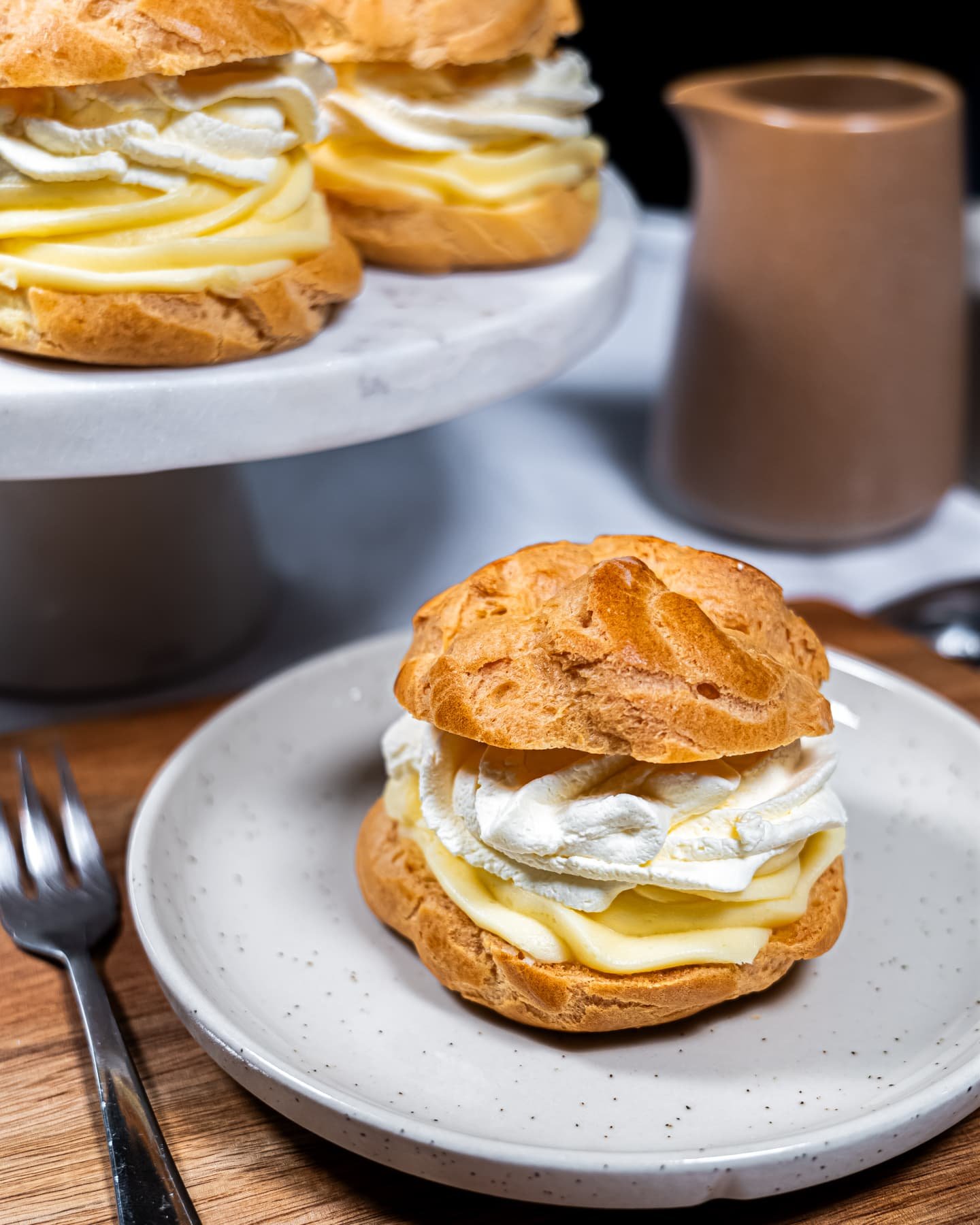 Golden cream puffs filled with silky vanilla pastry cream and fluffy whipped cream, served on a speckled ceramic plate with a cake stand in the background.