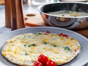 A freshly cooked egg white frittata with melted cheese, spinach, and bell peppers on a plate, with a steaming skillet in the background.