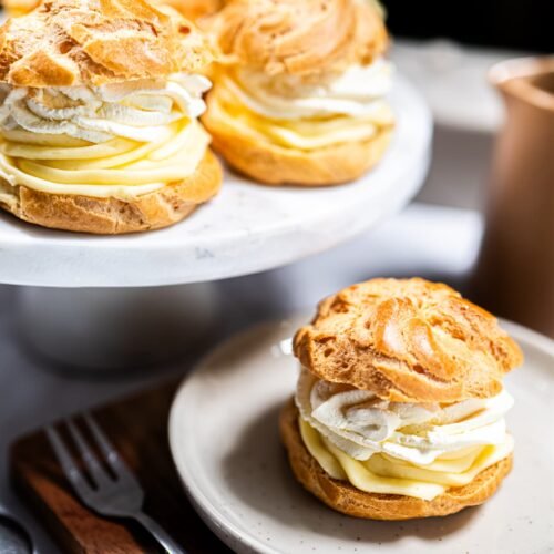 Golden-brown cream puffs filled with silky vanilla pastry cream and whipped cream, served on a marble cake stand and a rustic plate.
