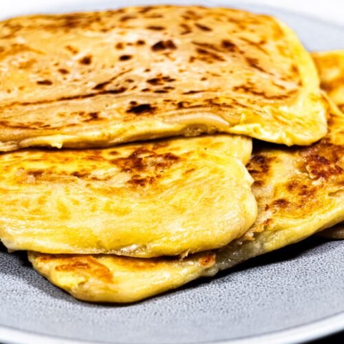 A stack of crispy, golden-brown Roti Prata filled with eggs and melted cheese, served on a plate.