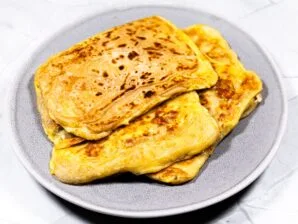 A plate of crispy, golden-brown Roti Prata filled with eggs and cheese, freshly cooked and ready to serve.
