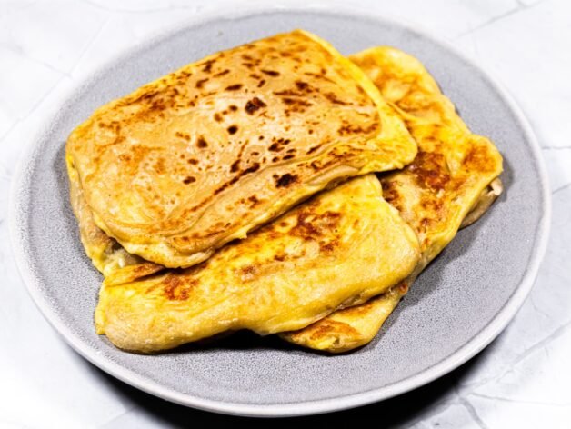 A plate of crispy, golden-brown Roti Prata filled with eggs and cheese, freshly cooked and ready to serve.