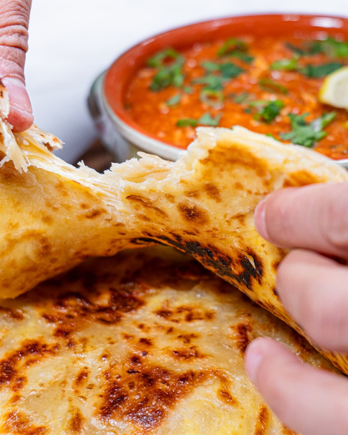 Hands tearing apart a crispy, flaky Roti Prata, revealing its soft layers, with a bowl of curry in the background.